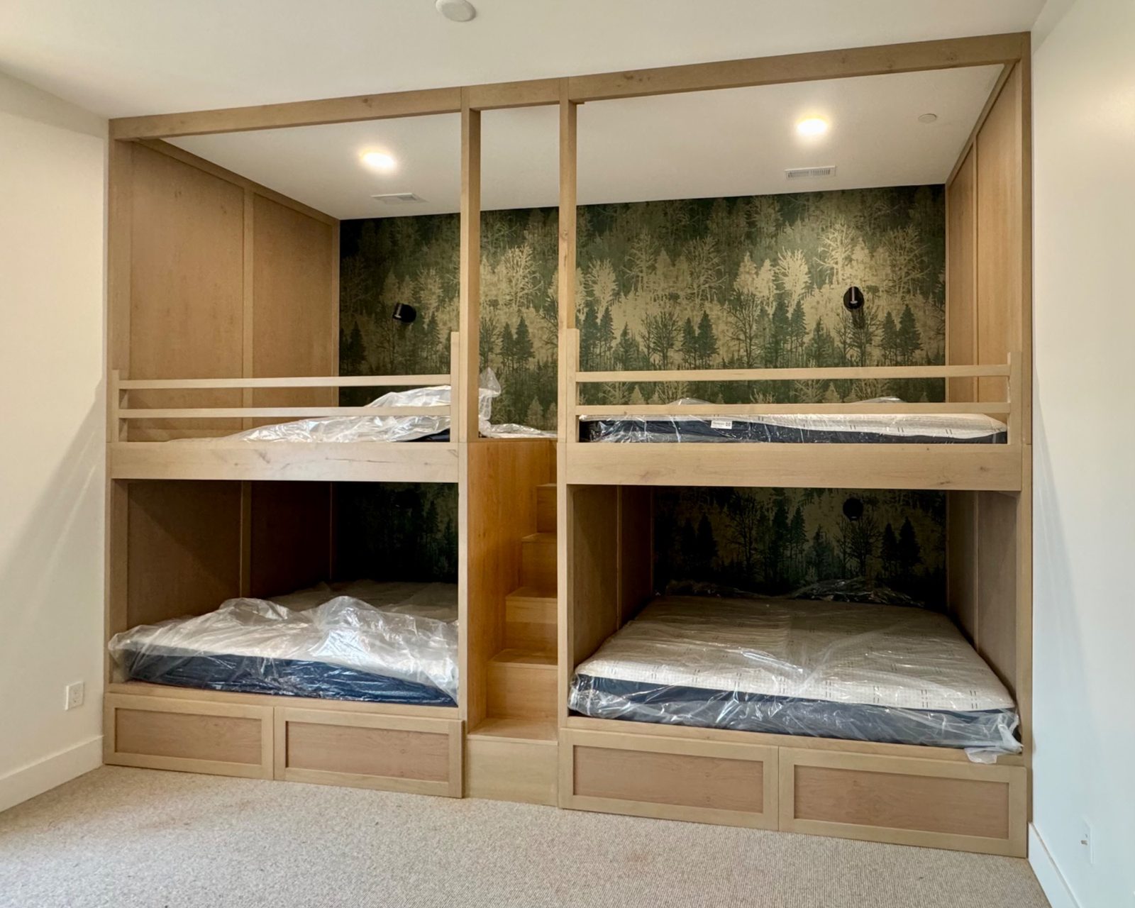 Goodman bunk room — four natural maple built-in bunks with forest wallpaper and center staircase